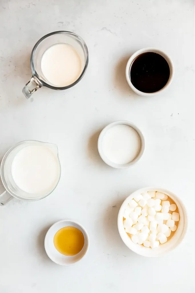 ingredients for homemade coffee creamer including marshmallows, half & half, heavy cream, vanilla pods, cinnamon sticks, honey, and chocolate syrup on wooden counter